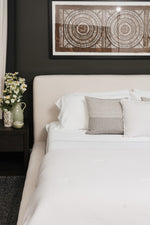 Smoothr Pearl White bamboo sheet set — A close-up of a neatly made bed with white bedding, a light beige upholstered headboard, and a textured gray accent pillow, set against a dark charcoal wall with framed circular artwork above.