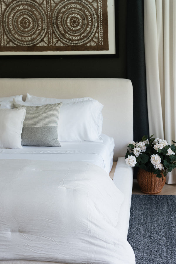 Smoothr Pearl White bamboo sheet set — A neatly made bed with white bedding and pillows, including a textured gray accent pillow, set in a modern bedroom with a cream upholstered headboard, dark wall, artwork, and a basket of white flowers beside it.