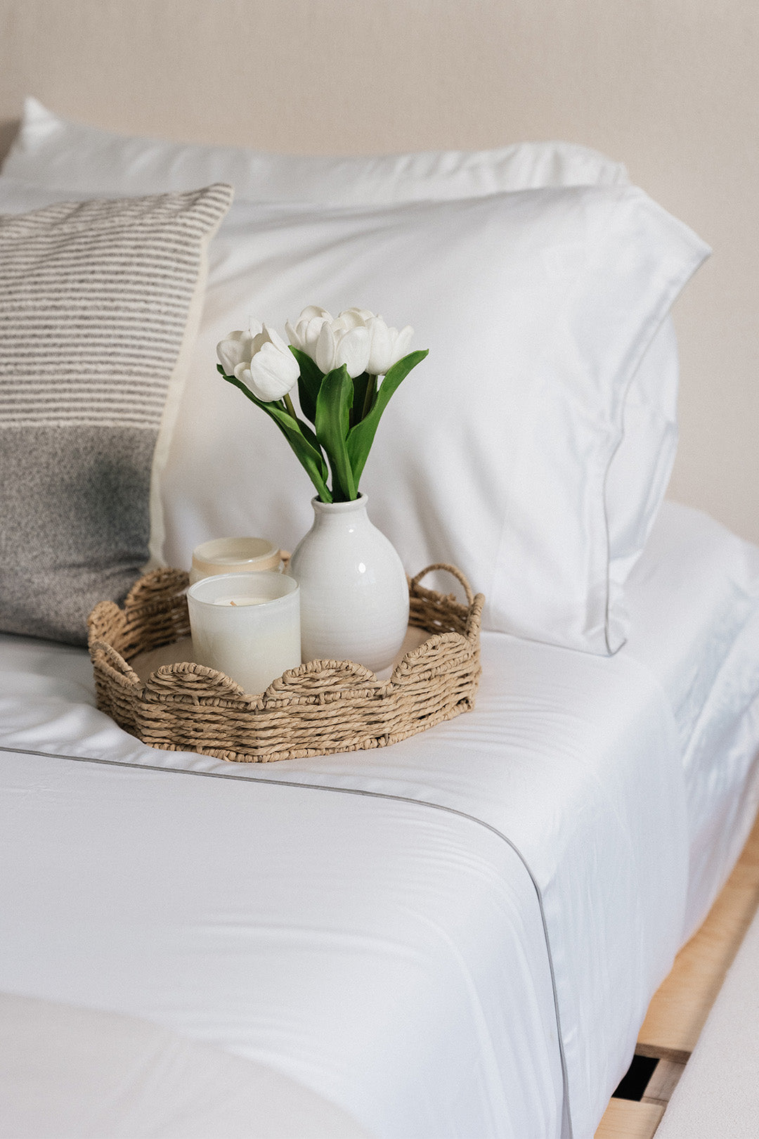 Smoothr Pearl White bamboo sheet set — A styled bedroom scene showing a white bed with pillows, a woven tray holding a white vase with white tulips and candles, and a gray textured accent pillow.