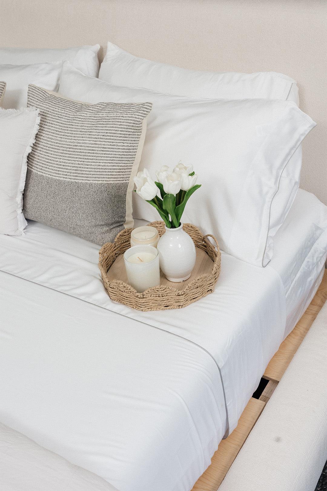 Smoothr Pearl White bamboo sheet set — A styled bed with white bedding, a textured gray accent pillow, and a woven tray holding white candles and a small vase of white tulips.