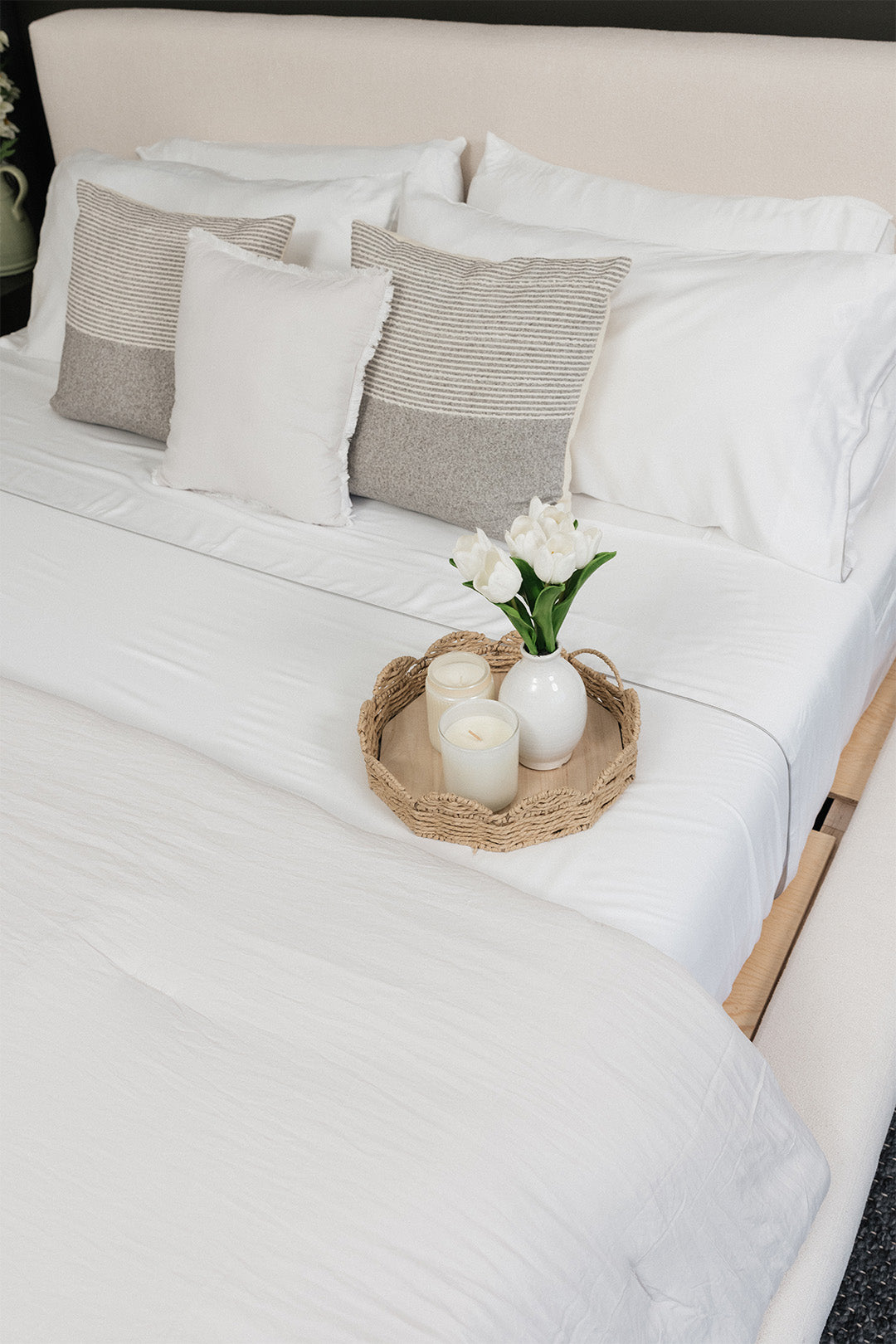 Smoothr Pearl White bamboo sheet set — A neatly made bed with white bedding, several decorative pillows in white and beige, and a woven tray holding white candles and a small vase of white flowers on the foot of the bed.