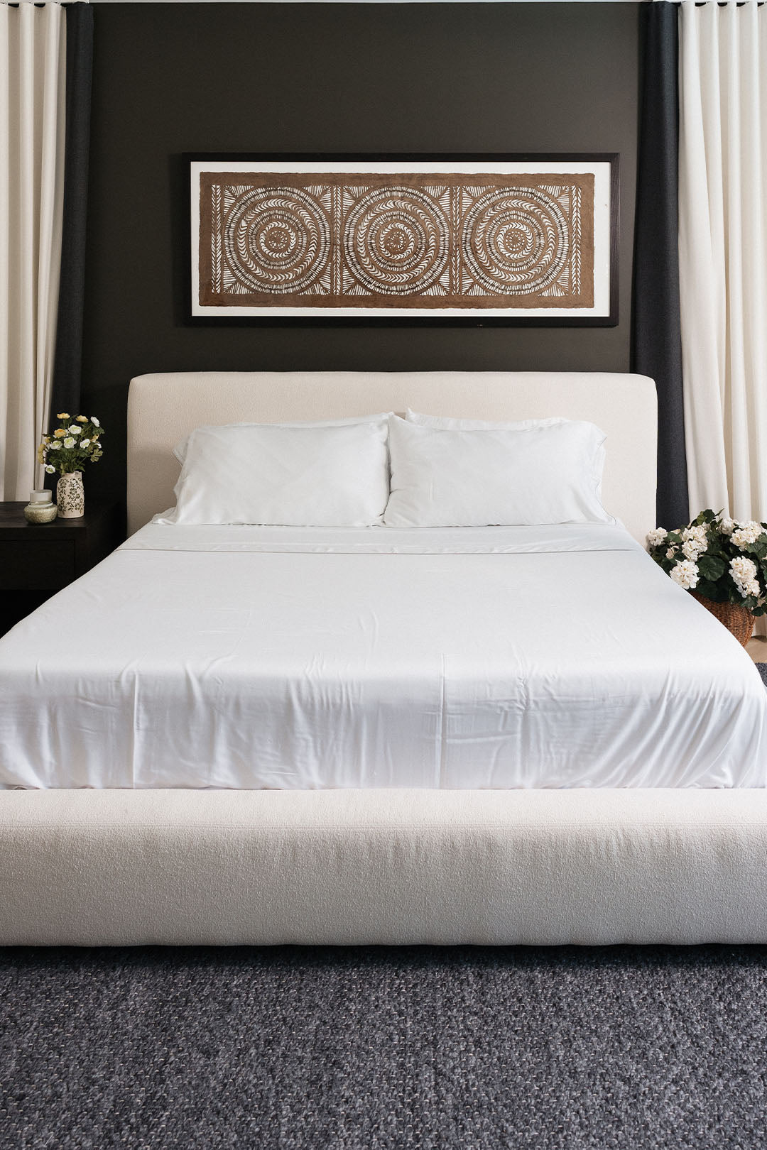 Smoothr Pearl White bamboo sheet set — A modern bedroom setup featuring a low-profile platform bed with a white upholstered headboard and matching white bedding. The bed is centered against a dark charcoal wall with a framed circular art piece hanging above.