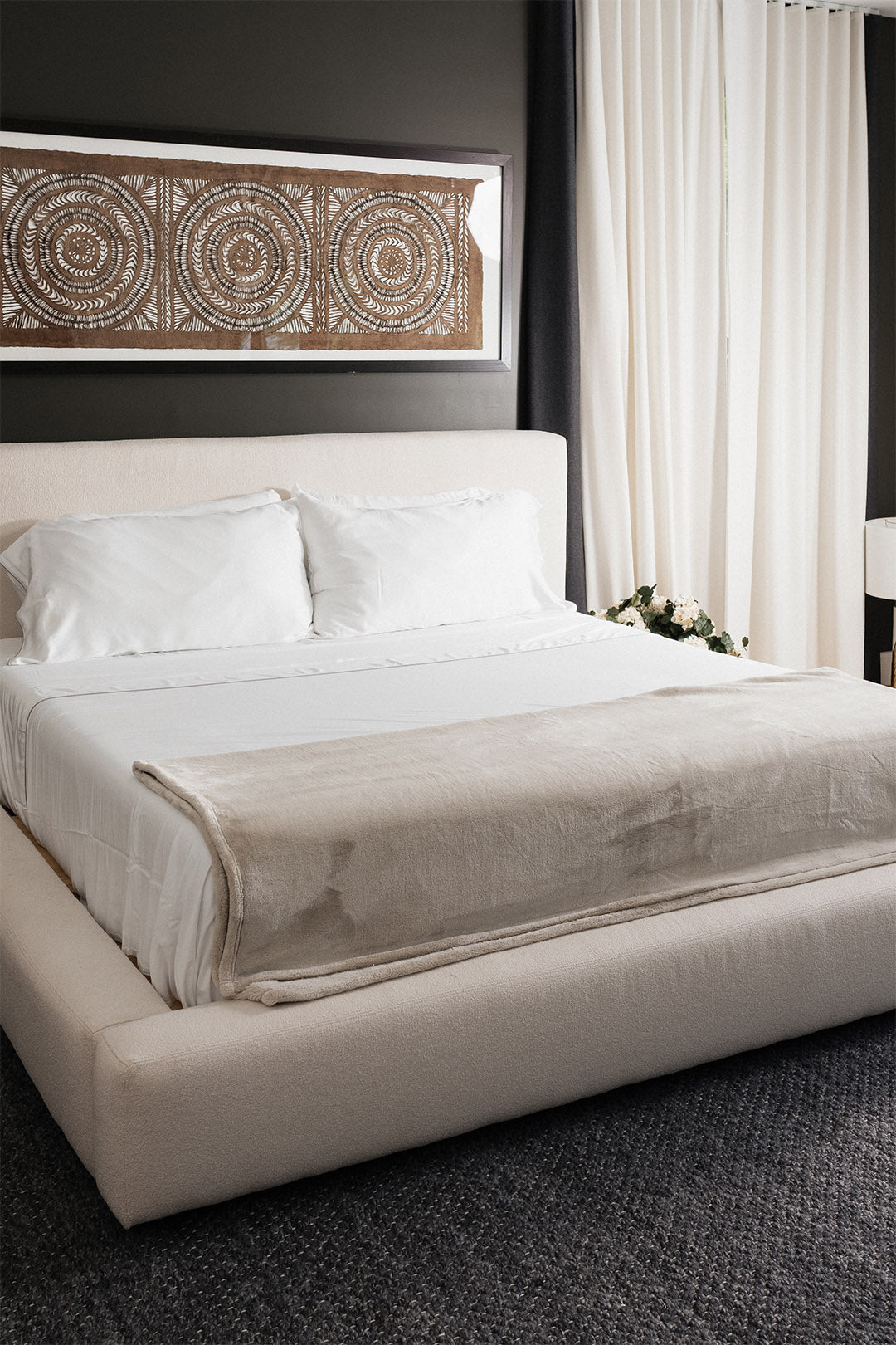 Smoothr Pearl White bamboo sheet set — A modern platform bed with a cream upholstered frame, dressed in white linens and a light beige throw blanket, set against a dark gray wall with a large framed mandala print above.